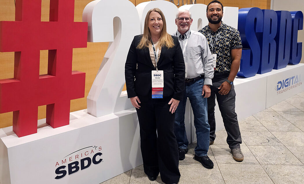 vicki, curt, and christopher are pictured standing in front of a large 3d display that reads hashtag twenty twenty four s b d c. they are smiling and wearing conference lanyards