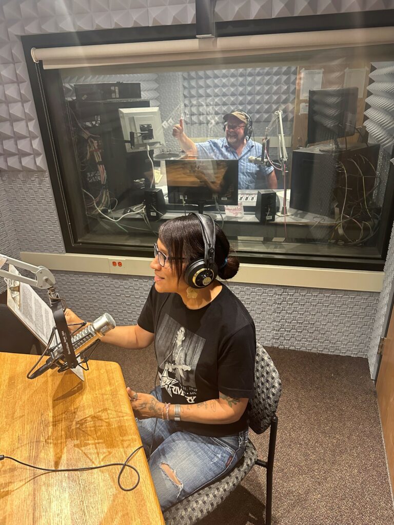 ivy vainio is seated in a recording studio wearing headphones with a microphone and script in front of her. a radio producer is behind glass in the booth in the background.