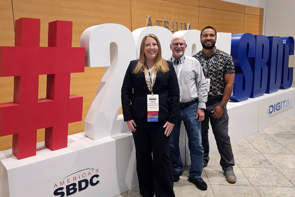 vicki hagberg, curt walczak, and christopher henagin are pictured standing in front of a large 3d hashtag display for america's small business development center 2024 national conference