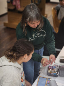 a young native american woman is seated at a work table and next to her stands sarah howes who is demonstrating a technique