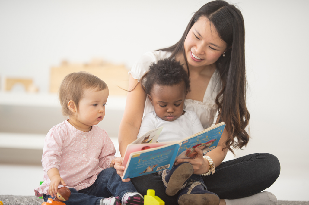 a female early childhood professional sits on the floor with two toddlers looking at a picture book